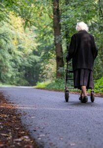 An elderly lady using a rollator to walk down a road in the woods