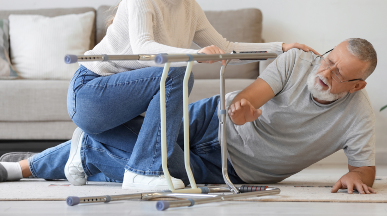 man with a walker fallen in living are with woman trying to help him up - How to Prevent Falls in Seniors