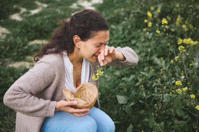 Young woman sneezing while gathering wildflowers in the meadow outdoors, suffering from allergy in the spring time - What are Seasonal Allergy Symptoms