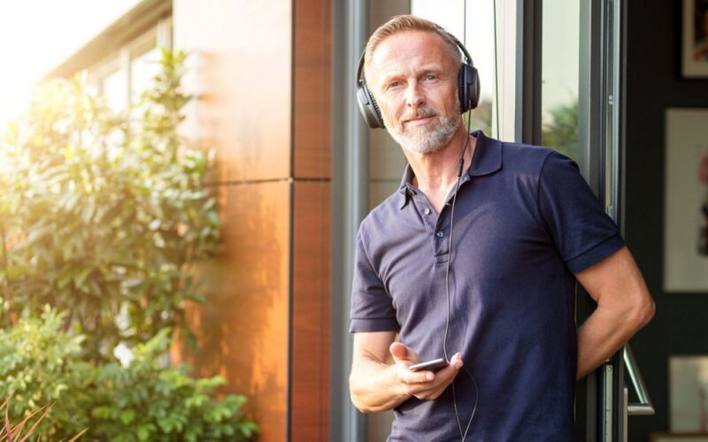 Senior standing against a door outside a building hold a cell phone in his hand connected to headphones - 6 Best Headphones for Seniors