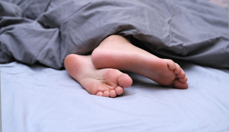 woman lying down on bed covered with a comforter with 2 feet showing - Restless leg Syndrome